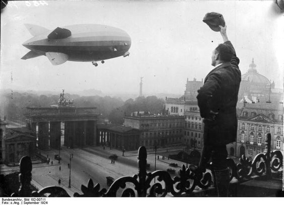 A black and white photograph of an airship flying past a landmark in 1920s Berlin, whilst an onlooker waves his cap.