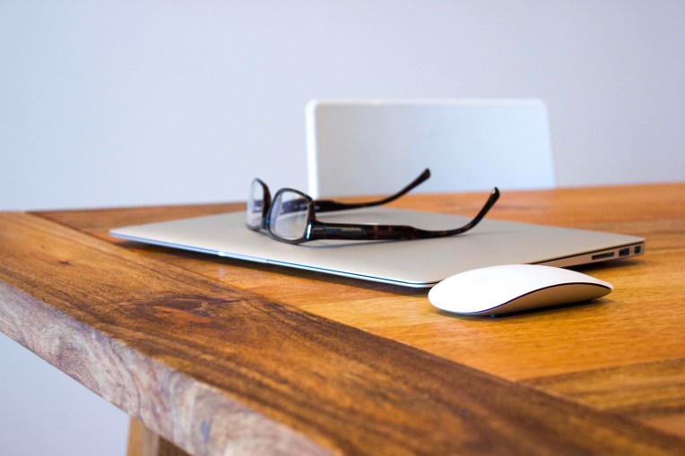 A closed laptop with a mouse and spectacles on a wooden table